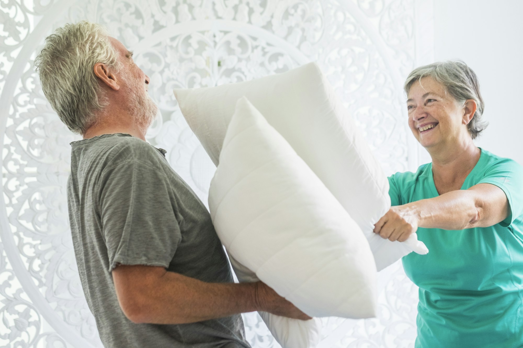 Happy old people couple play together in a bright room with war pillows having fun and enjoying