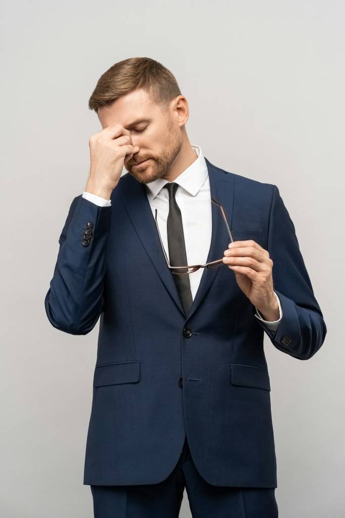 Frustrated businessman in emotional stress holds eyeglasses rubs bridge of nose on studio background